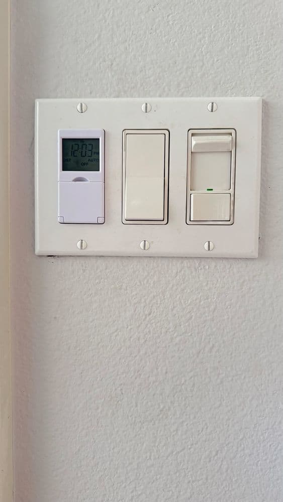 Digital thermostat and light switches on a white wall plate.
