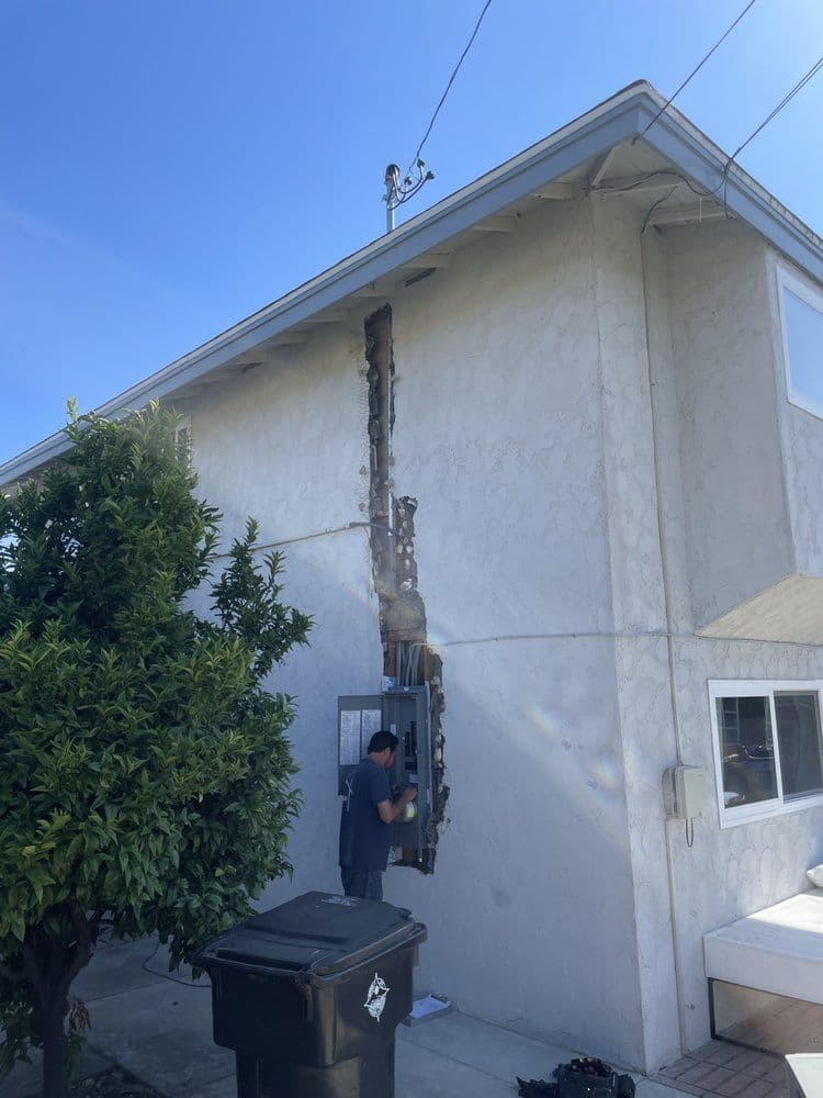 Repairing damaged exterior wall with exposed wood and electrical panel at residential property.