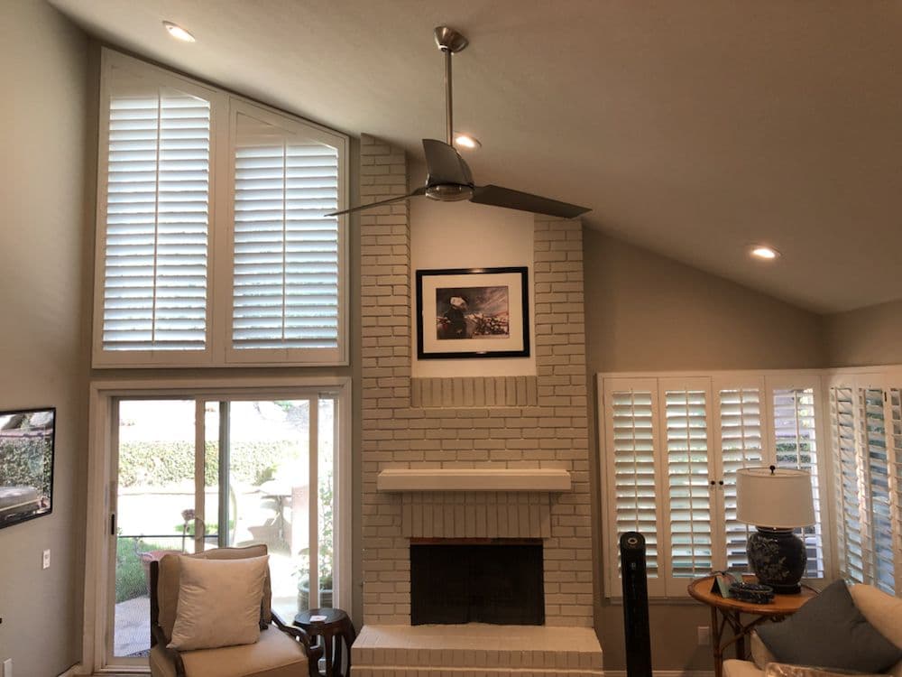 Bright living room featuring a white brick fireplace, modern ceiling fan, and large windows.