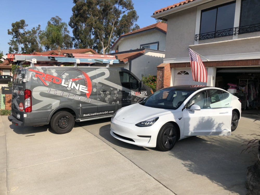 Red Line Electric van and white Tesla parked in residential driveway with American flag.