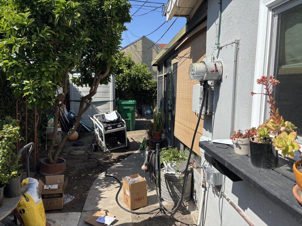 Garden pathway with potted plants, utility meters, and outdoor clutter in urban setting.