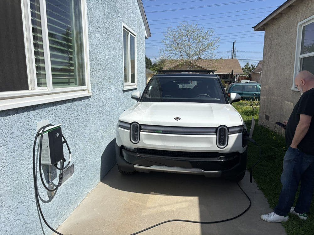 Electric vehicle charging at home beside a light blue house with a person nearby.