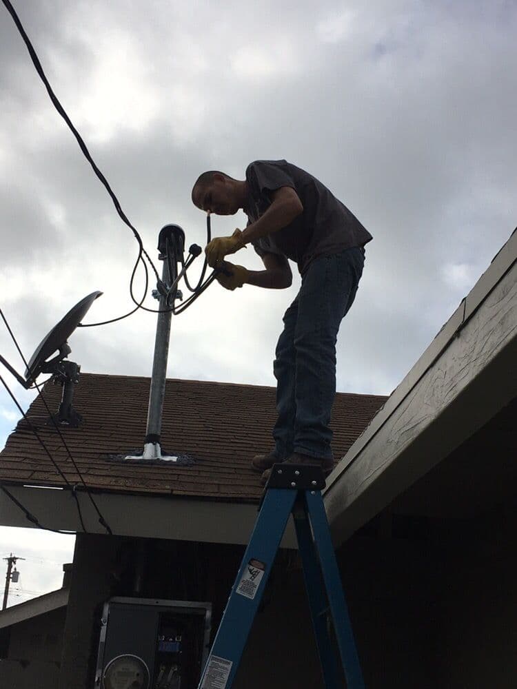 Technician adjusting satellite dish on rooftop using a ladder and safety gloves.