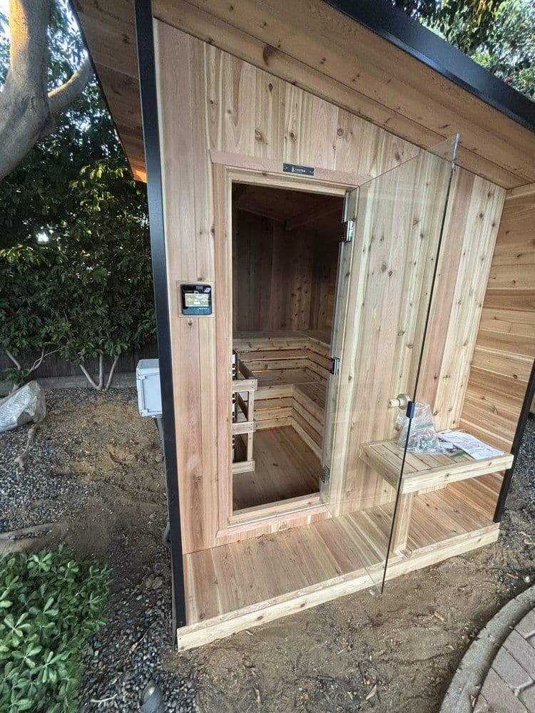 Modern wooden sauna with glass door in a backyard setting, featuring natural cedar interior.