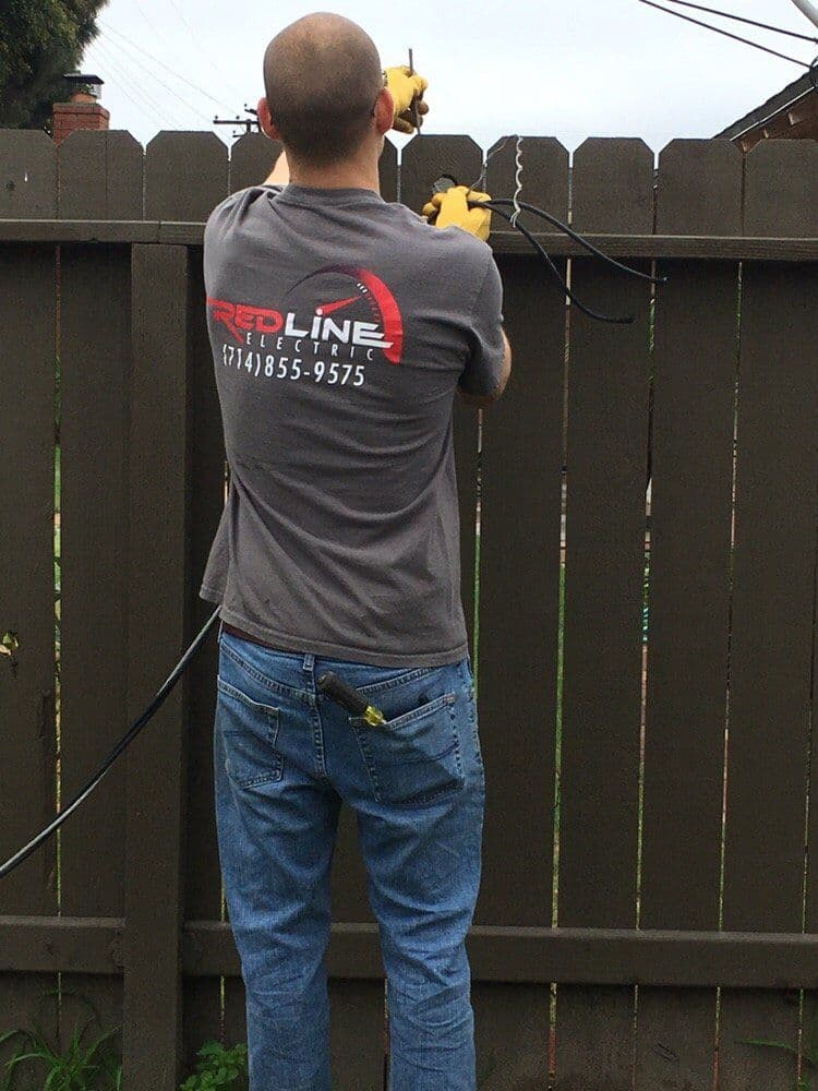 Electrician working on a fence with tools, wearing a Red Line Electric shirt.