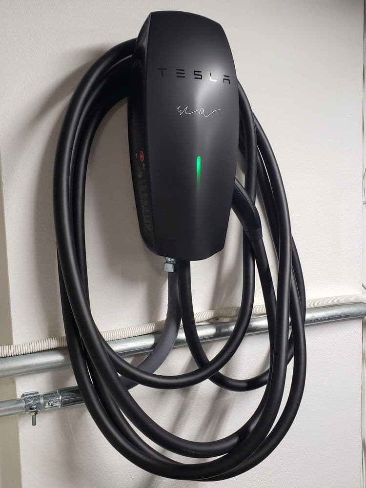 Tesla wall-mounted electric vehicle charger with coiled cable and green light indicator.