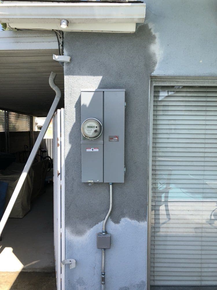 Electrical meter and panel mounted on a gray wall with nearby window blinds.