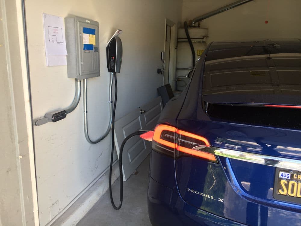 Blue Tesla Model X charging in a garage with electrical panel and wall-mounted charger.